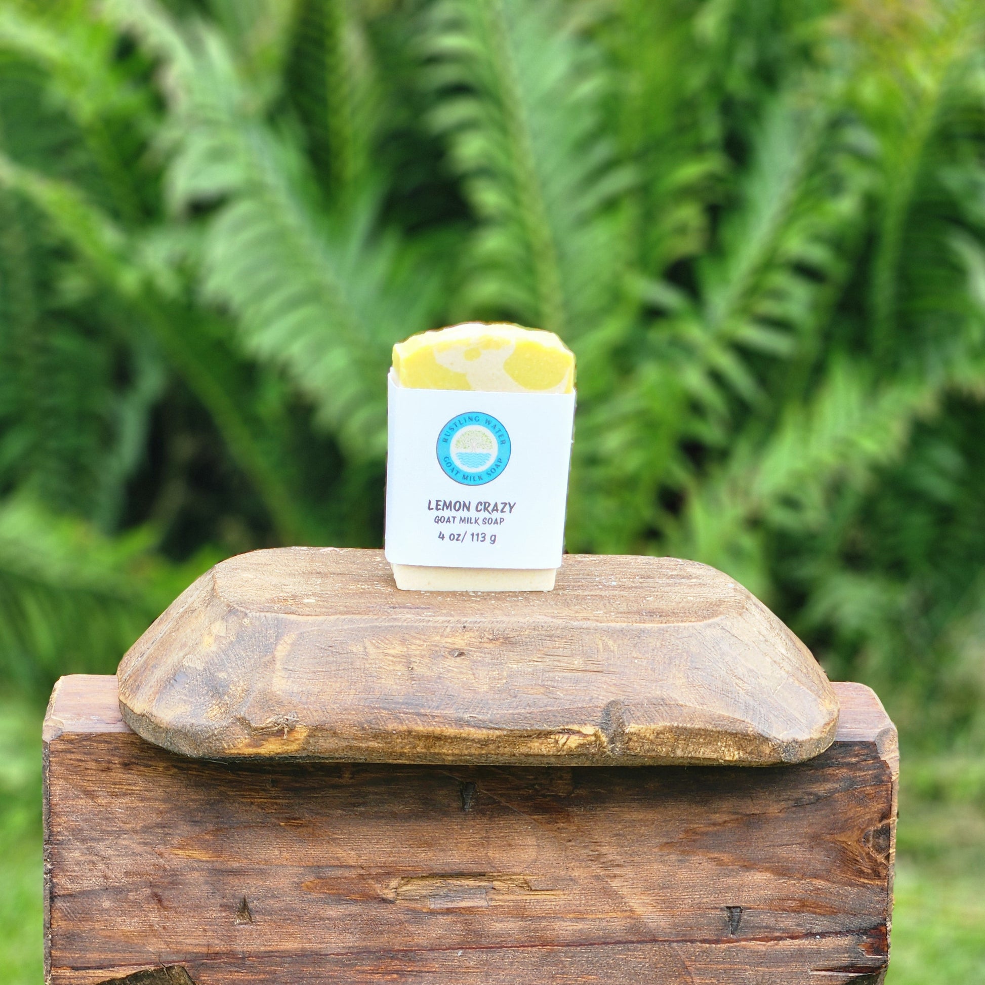 A small white soap bar labeled 'LEMON CRAZY' with yellow soap visible at the top, displayed on a rustic wooden box outdoors with a blurred green fern background.