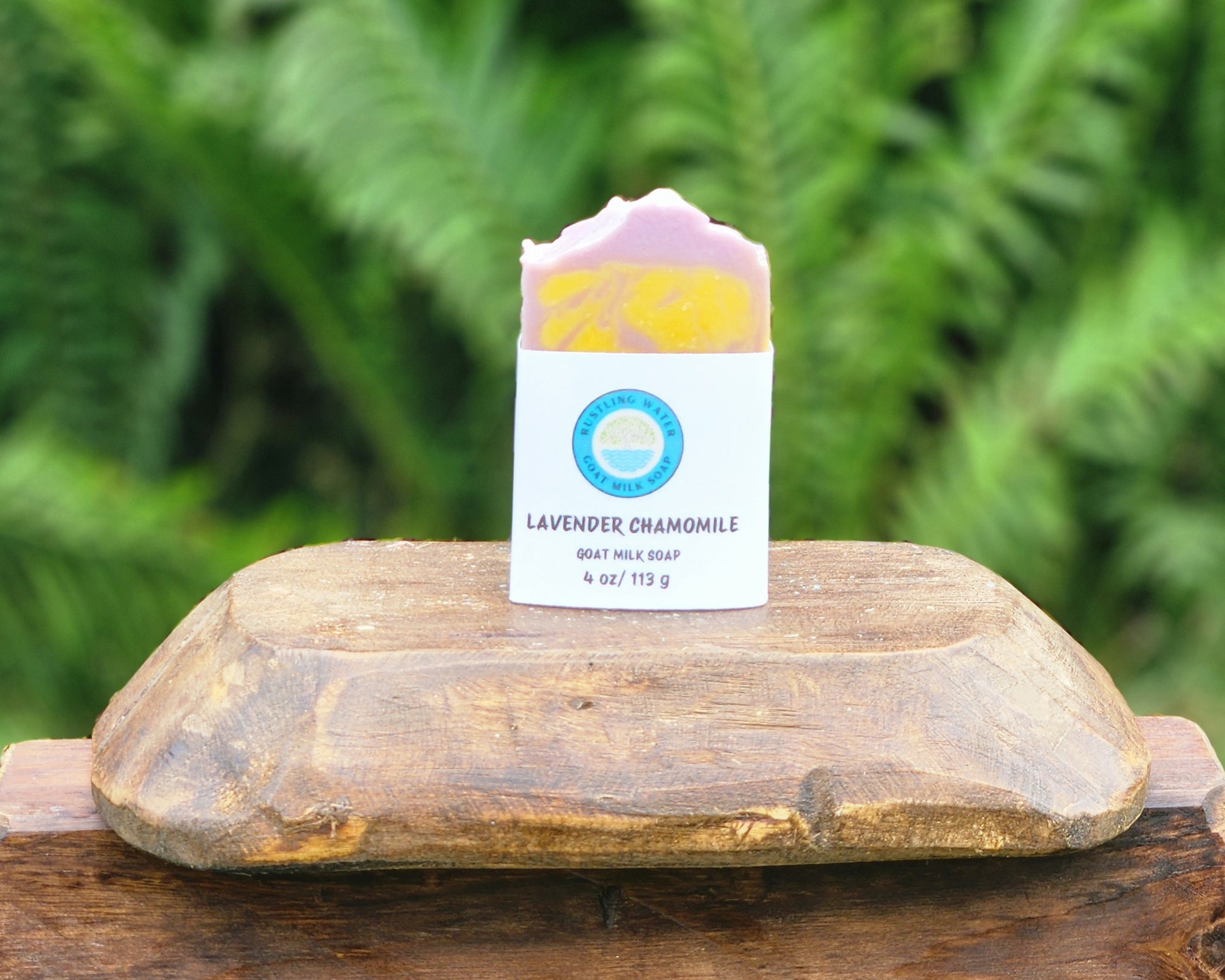 A handmade lavender chamomile soap bar in its white paper packaging, displayed on a rustic wooden box outdoors with a blurred green fern background.