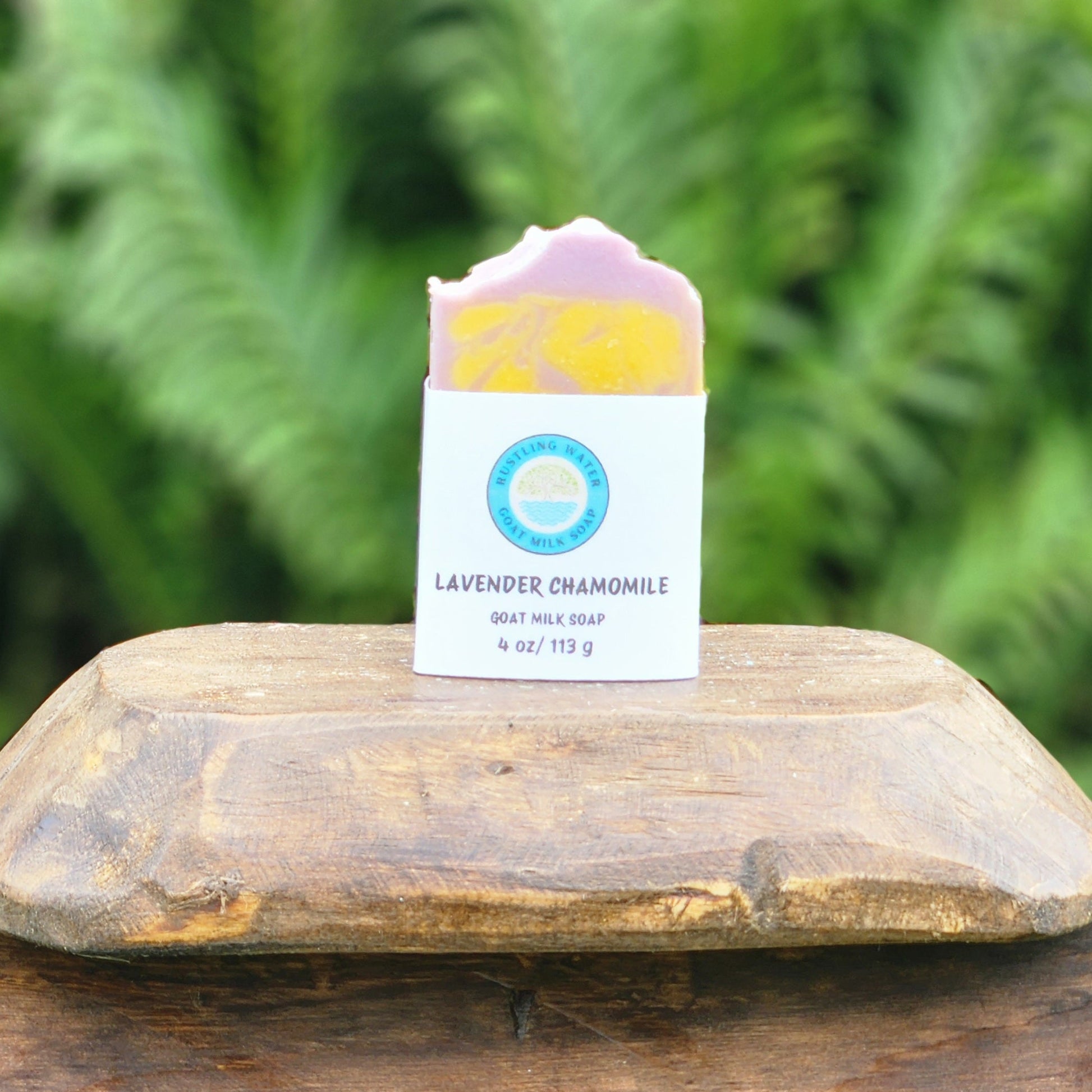 Lavender Chamomile hand soap in a white paper sleeve with a blue circular logo, resting on a rustic wooden box lid outdoors with a blurred green fern background.