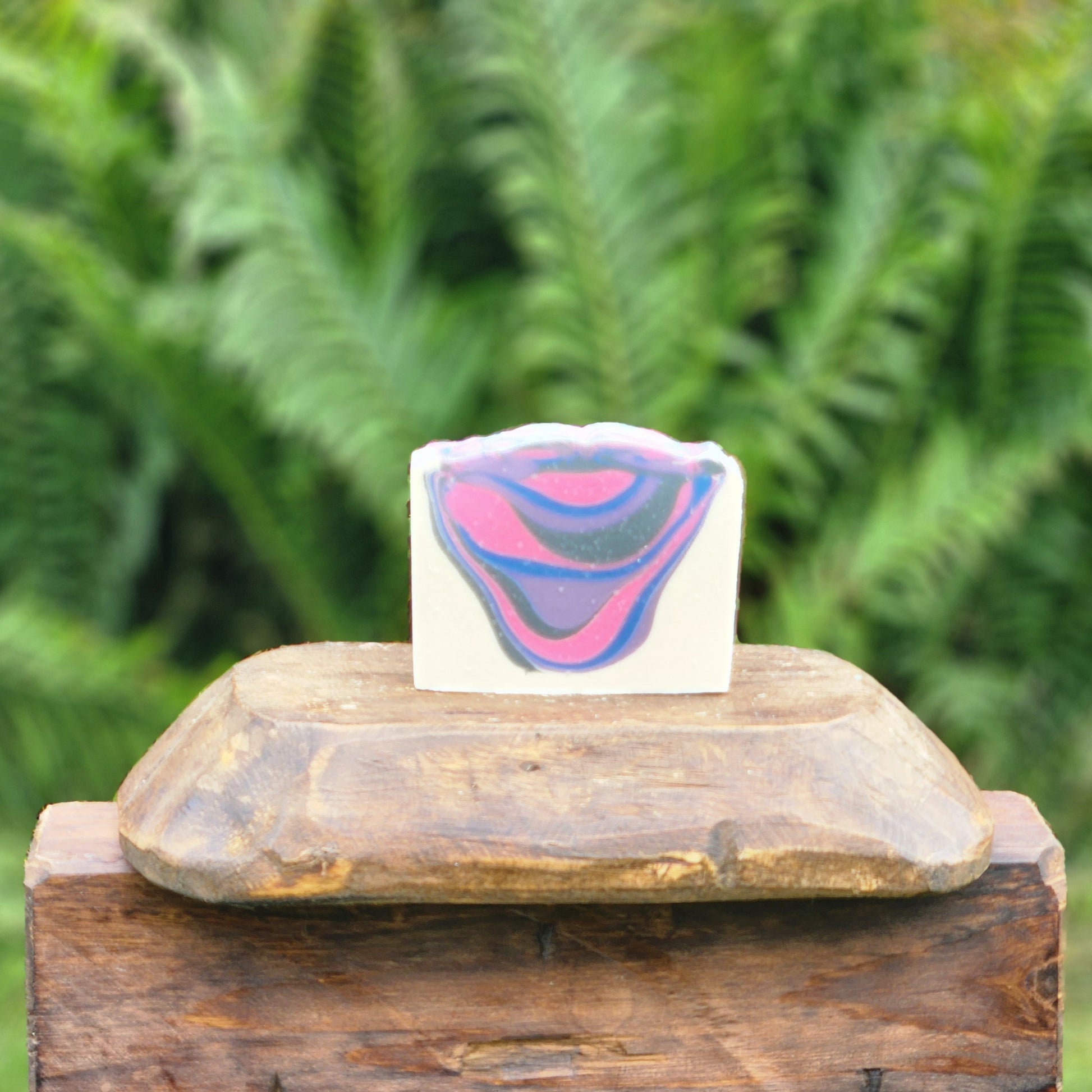 A rectangular handmade soap bar with a wavy, scalloped edge, featuring a marbled swirl pattern of pink, purple, and blue on a white base. The soap is displayed on a rustic wooden block with a blurred background of green ferns.