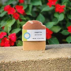 A rectangular bar of soap labeled 'FORGETTEN' stands on a stone ledge with a blurred background of red flowers and green leaves.