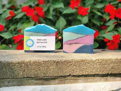 Two handcrafted rectangular bars of soap with a mountain-like layered design, displayed on a stone ledge with red flowers in the background. The left bar has a white label reading 'TIPSY LANE