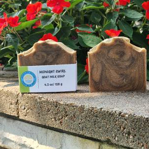 Two bars of Midnight Swirl goat milk soap displayed on a stone ledge with red flowers and green foliage in the background. One bar has a white paper label attached.