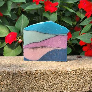 A rectangular bar of handmade soap with a wavy, layered pattern in shades of teal, light green, pink, and navy blue, resting on a rough stone ledge with red flowers and green leaves in the background.