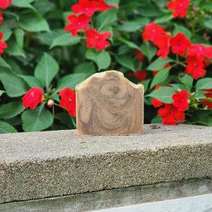 A rectangular bar of goat milk soap with a natural swirling wood grain pattern, standing upright on a concrete ledge with red flowers in the background.