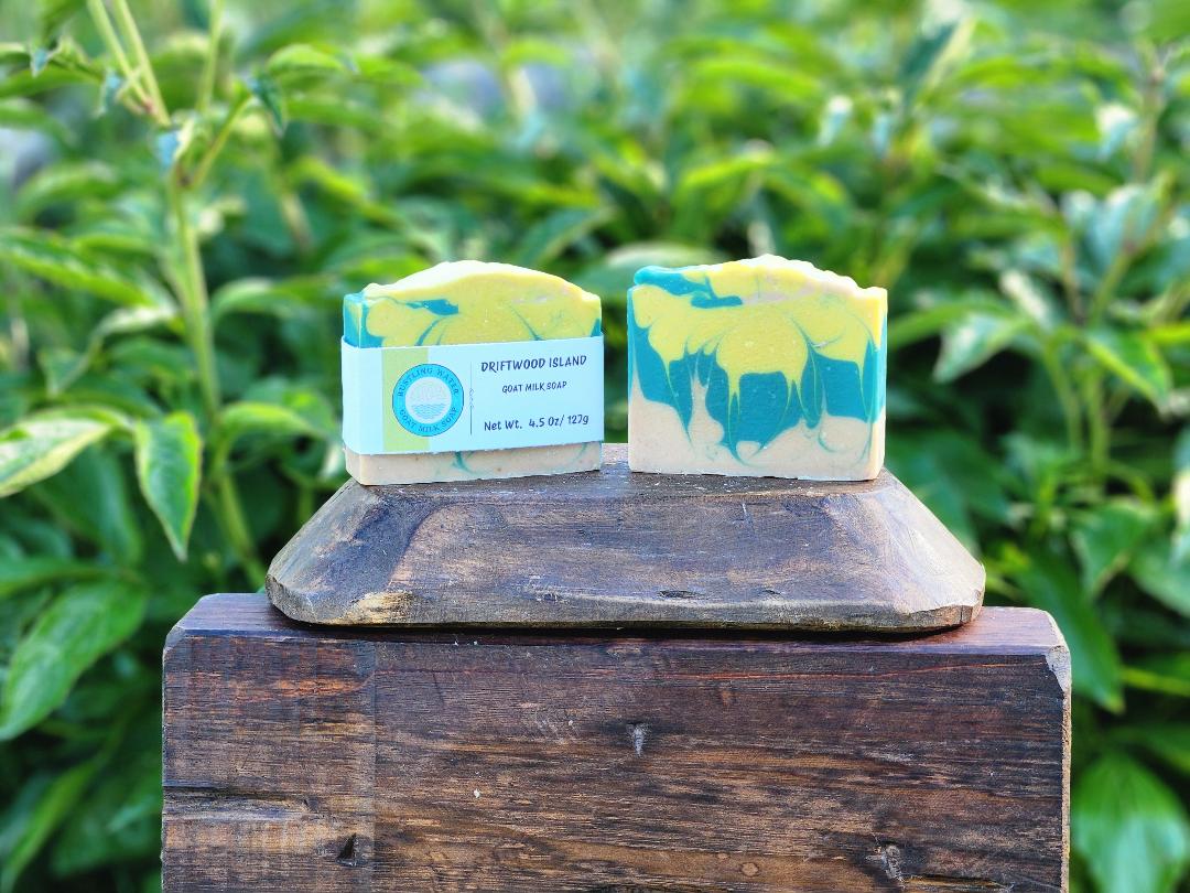 Two bars of soap colored yellow, blue and beige on a wooden display with a green leafy background.