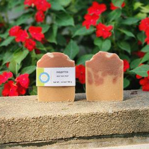 Two handmade soap bars in a warm tan-brown color, one with a paper label, displayed on a stone ledge with red flowers and green leaves in the background.